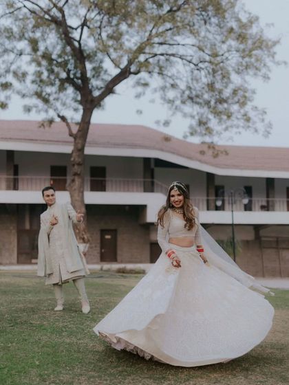 The bride twirls in her beautiful white lehenga while the groom looks on with a smile. A playful and happy moment.
