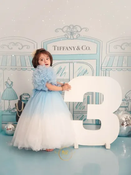 A three-year-old girl in a stunning blue gown, posing with a large number "3" during her chic "Tiffany & Co." themed birthday shoot.