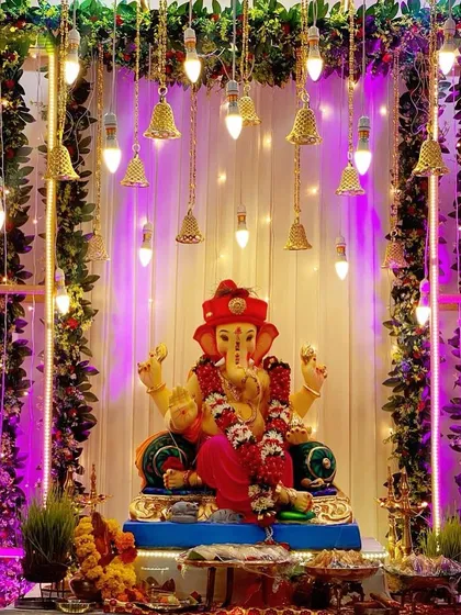 A beautiful Ganpati mandap featuring a canopy of green vines and hanging lighted bells. The purple lighting creates a magical and devotional atmosphere.