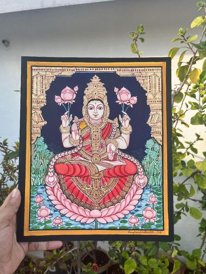 A compact yet divine 10x12 inch painting of Goddess Lakshmi. The dark background makes her form, adorned with gold and seated on a pink lotus, truly shine.