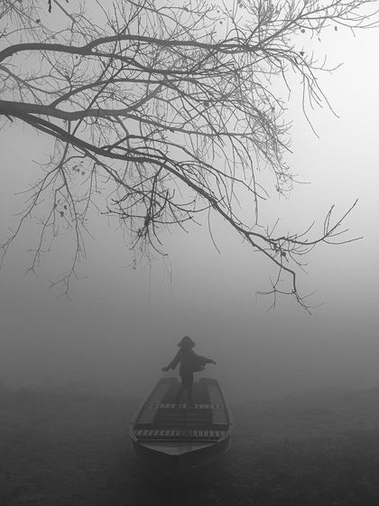 A figure stands in a boat, enveloped by thick fog and framed by the skeletal branches of a tree. This black and white image is the essence of peace and solitude.