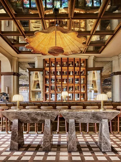 The bar at One8 Commune in Hyderabad is anchored by monolithic marble tables, creating a powerful statement of contemporary luxury. Above, a massive rattan light fixture and a mirrored ceiling amplify the sense of scale and tropical maximalism that defines the restaurant's character.