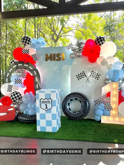 An outdoor Cars-themed first birthday party. The setup includes a Lightning McQueen cutout, a light-up number '1', and a Route 1 themed plinth.