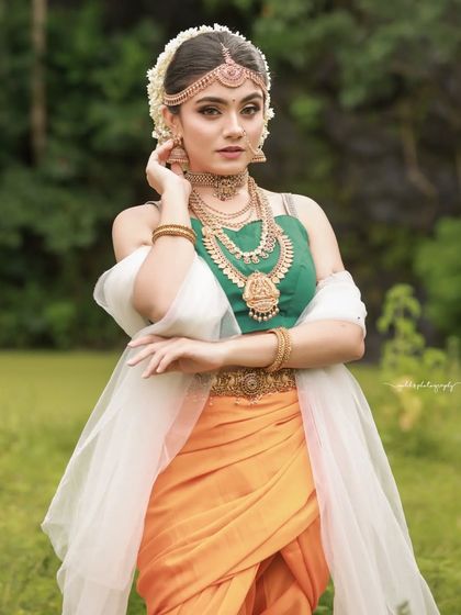 Inspired by the grandeur of Bahubali, this "Sakhi" look features a model in a unique orange and green dhoti-style saree. The outdoor setting and regal jewelry complete the ancient princess aesthetic.