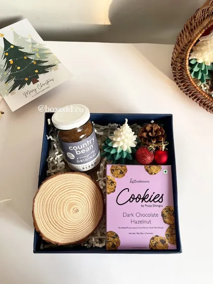 A cozy Christmas-themed gift box with dark chocolate hazelnut cookies, artisanal coffee, a wooden coaster, and festive tree-shaped candles.
