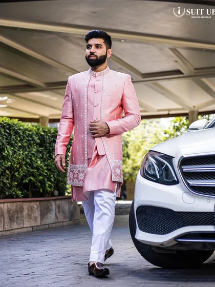A client looking dapper in a custom pink Indo-Western set. The jacket features an asymmetrical cut with delicate embroidery on the hem and placket, paired with a matching kurta and white trousers for a fresh, modern wedding look.