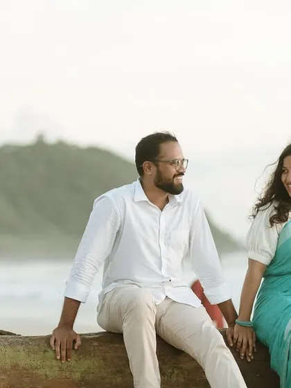 The couple sitting on a rustic log by the sea, sharing a laugh against the beautiful backdrop of the coastline and hills.