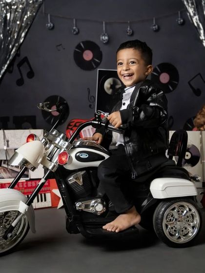 This toddler is all smiles in a rock-and-roll biker themed shoot. Dressed in a leather jacket, he sits on a toy motorcycle against a backdrop of vinyl records and silver streamers.