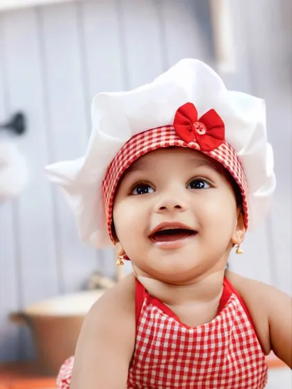 A joyful close-up of our future master chef, smiling brightly in her adorable red-and-white checkered apron and hat.