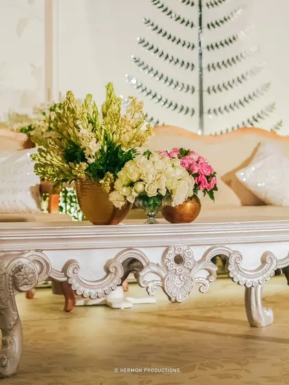 A close-up of an intricately carved white coffee table, topped with elegant floral arrangements in golden vases. The craftsmanship of the furniture is a key element of the luxurious design.