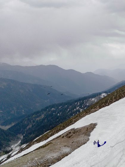 A breathtaking wide shot of the couple, a small blue speck in the vast, snow-covered mountain range.