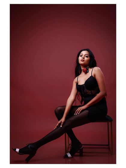 A glamorous and dramatic studio shot. The model is seated in a black lingerie-style top and tights against a rich red background, creating a look that is both seductive and high-fashion.