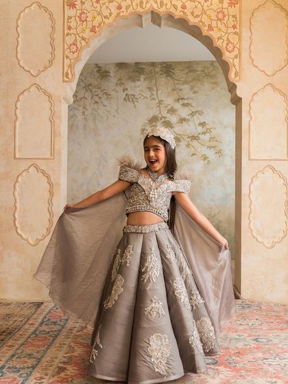 This stunning grey lehenga features off shoulder feather details and intricate silver embroidery, complete with a flowing cape.