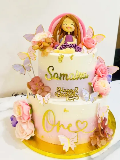 A dreamy first birthday cake with a beautiful fondant doll, rainbows, and butterflies. The pastel colors create a soft and magical feel.