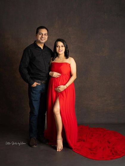 A full-length portrait of a couple in a classic studio setting. The mother-to-be looks radiant in a flowing red strapless gown, standing proudly with her partner against a dark, textured backdrop.
