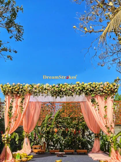 A daytime mandap setup with peach drapes and a floral canopy, set against a brilliant blue sky, perfect for a morning summer wedding.