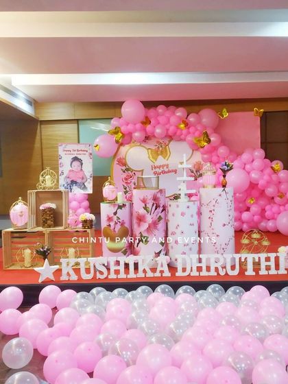 An elaborate first birthday decoration for a baby girl in shades of pink. The stage is filled with balloons, floral-printed pedestals, and a large 'ONE' sign.