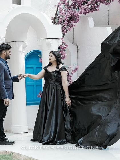 A couple holds hands against a white architectural backdrop, the bride's black trail gown billowing dramatically, creating a high-contrast, artistic photo.