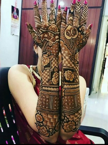 A bride showcasing her full bridal mehndi, which includes floral patterns, checkerboard fills, and a heart with the couple's initials, "SD".
