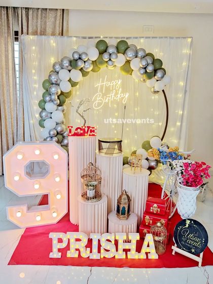 A chic birthday setup with an olive green, white, and silver balloon arch. The decor includes a large marquee number '9' and a neon sign.