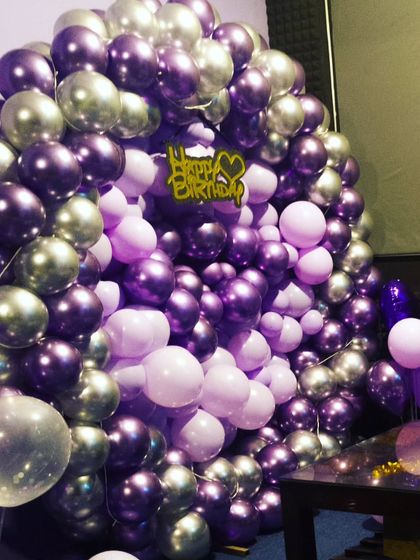 A dense wall of purple and silver balloons creates a rich, textured backdrop for a birthday celebration. The golden 'Happy Birthday' topper adds a nice finishing touch.