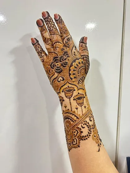 This intricate design features delicate 'jhumka' or earring motifs hanging from the wristband. It's a beautiful example of how we can incorporate jewelry elements into henna art.