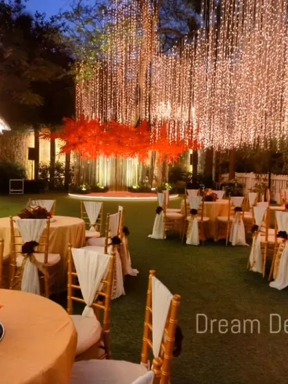 A wide view of an outdoor reception with a warm, autumnal color palette. The tables are set for guests, leading to a stage featuring red trees under a canopy of hanging lights.