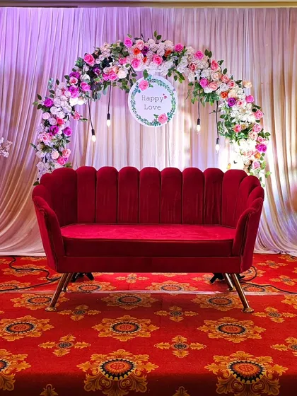 A wider view of the engagement stage decor. The floral arch and elegant red sofa are flanked by matching flower arrangements on pedestals, creating a balanced and formal look.
