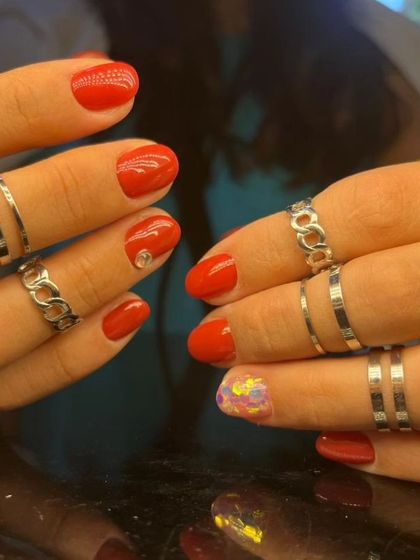 A vibrant red manicure with a single rhinestone accent and a fun, multi-colored confetti accent nail.