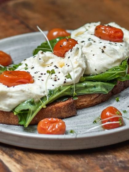 A beautiful plate of our Burrata and Confit Tomato Toast. Creamy, fresh burrata is spread over toasted sourdough and topped with sweet, slow-roasted cherry tomatoes.