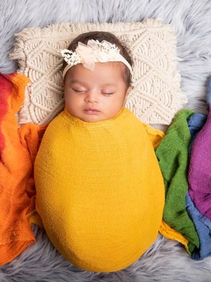 A rainbow of colors surrounds this peacefully sleeping newborn. The baby is wrapped in yellow, with colorful cloths fanning out, creating a bright and joyful portrait on a soft, furry rug.