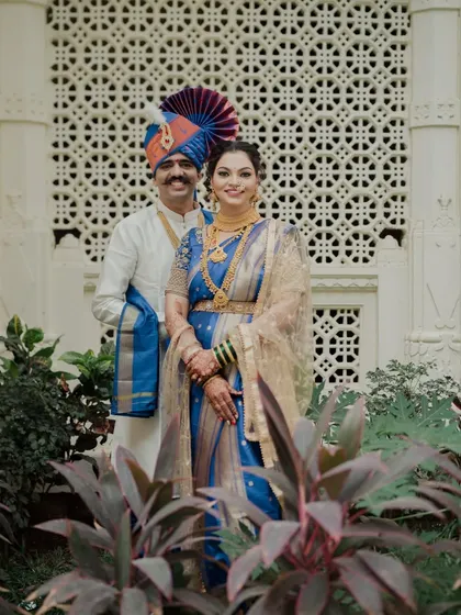 A full-length portrait of the couple set against an intricately carved wall. The lush greenery in the foreground adds a layer of depth, creating a rich and beautifully composed image.