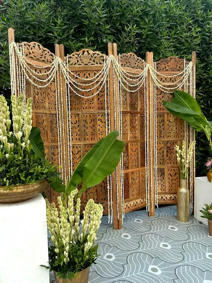 A carved wooden screen draped with jasmine strings, paired with tuberoses and banana leaves, creates an elegant and traditional partition or backdrop.