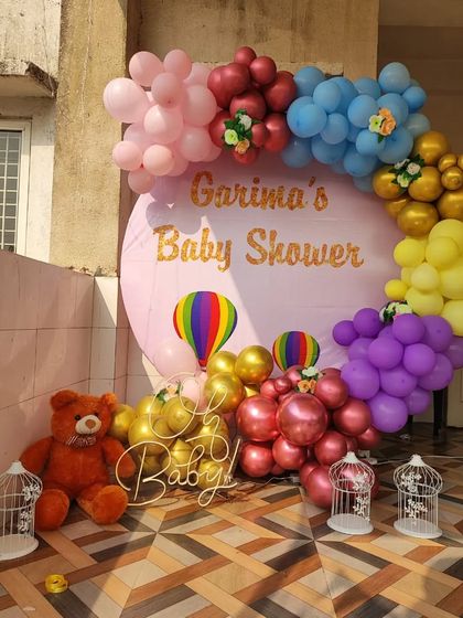 A colorful and cheerful baby shower backdrop for Garima. This outdoor setup features a rainbow-themed balloon arch around a personalized pink backdrop, complete with a teddy bear and other cute props.