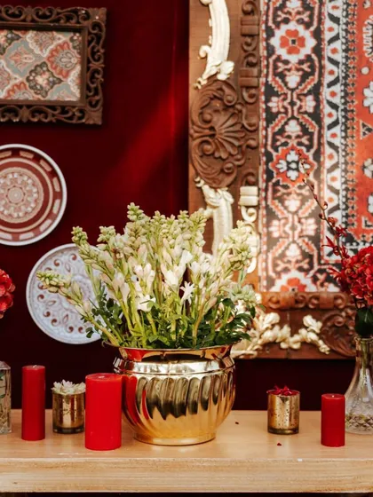 A beautiful arrangement of white tuberoses in a brass pot, placed against a backdrop of carved wooden panels and decorative plates. This highlights the blend of floral beauty and artisanal details.