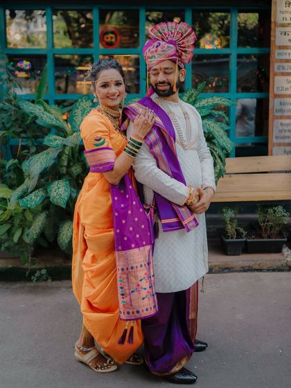 Rahul and Jagruti in a stylish pose, showcasing their vibrant orange and purple traditional outfits. The bride's navari drape is beautifully featured.