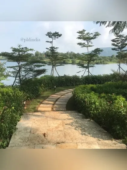 A stone pathway leads down to a serene lake. The path is lined with hedges and punctuated by Bucida trees, creating a beautiful, shaded walk to the water's edge.