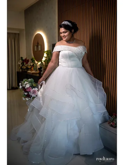 A full-length shot of the bride looking radiant in her off-shoulder wedding gown, holding her bouquet. This photo captures her happy glow and the beautiful details of her dress.