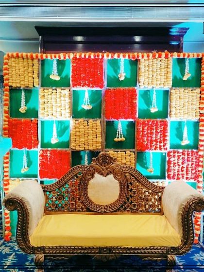 A festive backdrop featuring a checkerboard pattern of red, green, and woven straw panels, with hanging flower tassels. This design is perfect for a baby shower or Haldi ceremony, bringing texture and bright colors to the celebration.