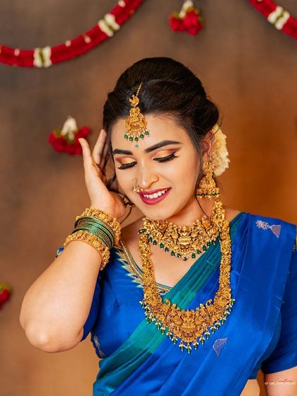 A bright and joyful close-up of a model in a brilliant blue saree. Her smile and pose highlight the matching temple jewelry set, including the necklace, earrings, and bangles, in a warm, inviting light.