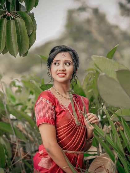A candid shot of a bride in a red saree, surrounded by lush greenery.