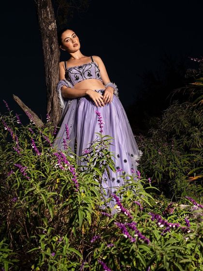 A full-length portrait that captures the model leaning against a tree, surrounded by flowers. The composition is dramatic and beautifully lit, like a scene from a dream.