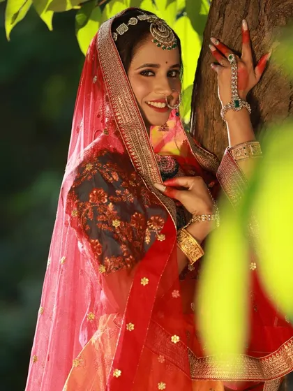 Framing the shot with natural elements like leaves adds depth and context. This portrait captures the model in a classic pose, highlighting the intricate jewellery and the rich red dupatta.