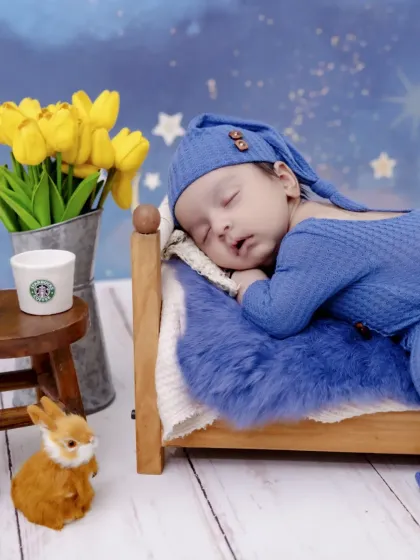 Dreaming under a starry night. This newborn is peacefully asleep in a miniature bed, dressed in a cozy blue outfit, with a whimsical backdrop of stars and moons for a magical bedtime story theme.