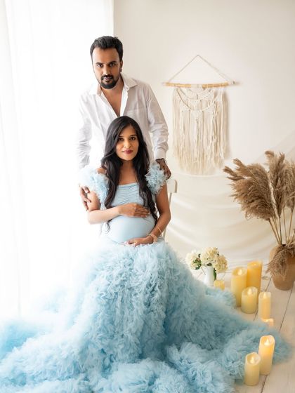 A beautiful portrait of the couple together. She is seated in the flowing blue gown while he stands behind her, both looking towards the camera, ready to welcome their little one.