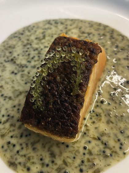 A close-up of pan-seared salmon with perfectly crispy skin. It's served on a bed of creamy sauce and garnished with delicate finger lime caviar.