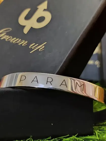 A silver cuff engraved with 'PARAM' sits inside its black 'Grown up' branded gift box, ready for gifting.