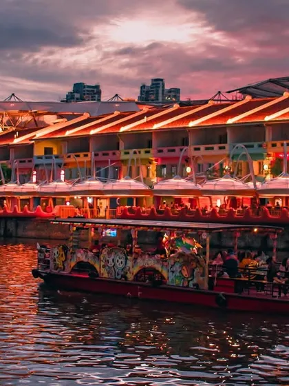 The colorful shophouses of Clarke Quay in Singapore are aglow with lights at dusk, as a river cruise boat passes by on the water.