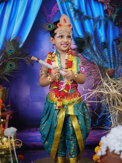 A smiling 'Little Krishna' holding his flute. The vibrant blue background and peacock feathers complete this beautiful and festive traditional portrait.