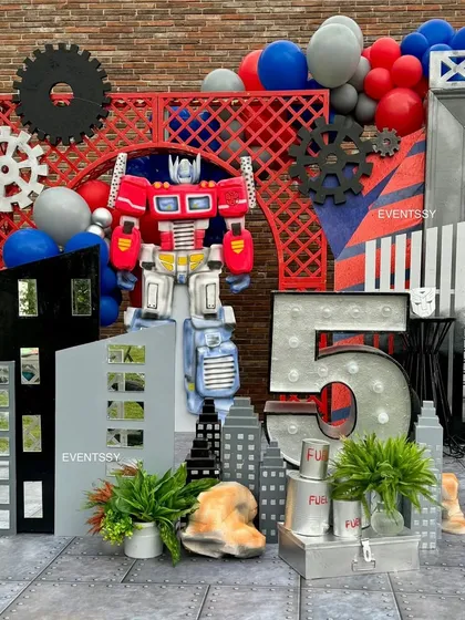 A focused view of the Optimus Prime centerpiece, set against a red industrial-style backdrop with gear props. The large marquee number "5" clearly marks the milestone birthday.